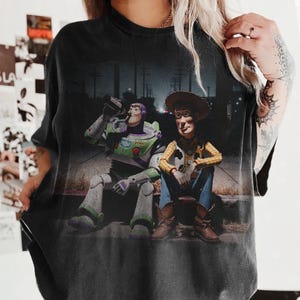 May include: Black t-shirt with a graphic of Buzz Lightyear and Woody from Toy Story. Buzz is drinking from a bottle. Woody is wearing a cowboy hat, yellow shirt, blue jeans, and brown boots.