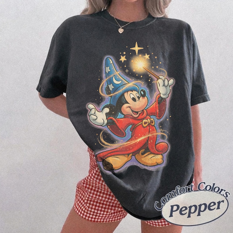 Pu&ograve; includere: T-shirt grigio scuro con un design di Topolino dei cartoni animati vestito da mago, che tiene in mano una bacchetta con effetti scintillanti. Il design include un cappello da mago blu con stelle e una veste rossa. La maglietta ha il testo "Comfort Colors Pepper" in un ovale bianco.