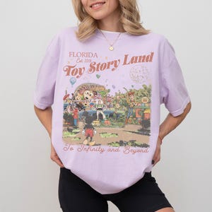 Peut inclure: Un t-shirt lilas avec un graphisme color&eacute; de Toy Story Land en Floride. Le design comprend Mickey Mouse, Woody et d'autres personnages, avec le texte "Toy Story Land" et "To Infinity and Beyond."