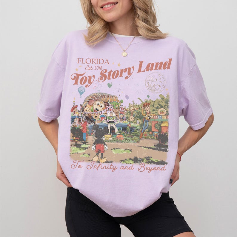 Peut inclure: Un t-shirt lilas avec un graphisme color&eacute; de Toy Story Land en Floride. Le design comprend Mickey Mouse, Woody et d'autres personnages, avec le texte "Toy Story Land" et "To Infinity and Beyond."