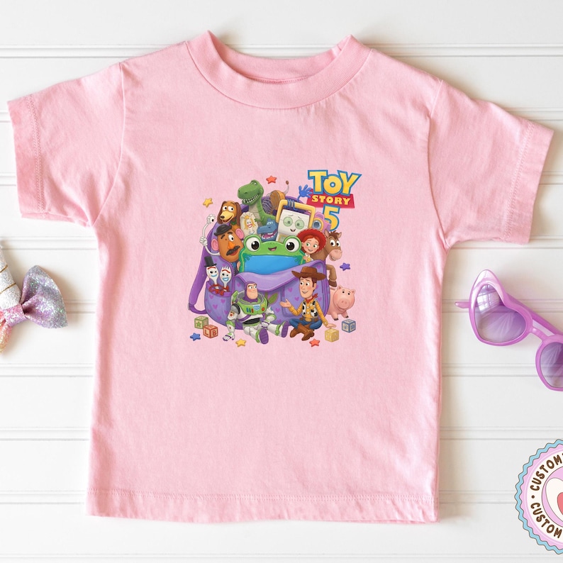 May include: A light pink short-sleeve t-shirt featuring a central graphic print of various Toy Story characters emerging from a purple backpack, with the text Toy Story 5 displayed in blue and yellow letters above the character group.