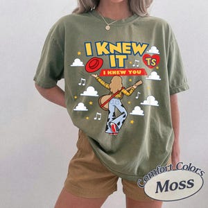May include: An olive green t-shirt featuring a graphic print with the text I KNEW IT I KNEW YOU and TS inside a red heart. The design includes a cartoon figure with blonde hair wearing a yellow plaid shirt and cow print trousers, holding a guitar.