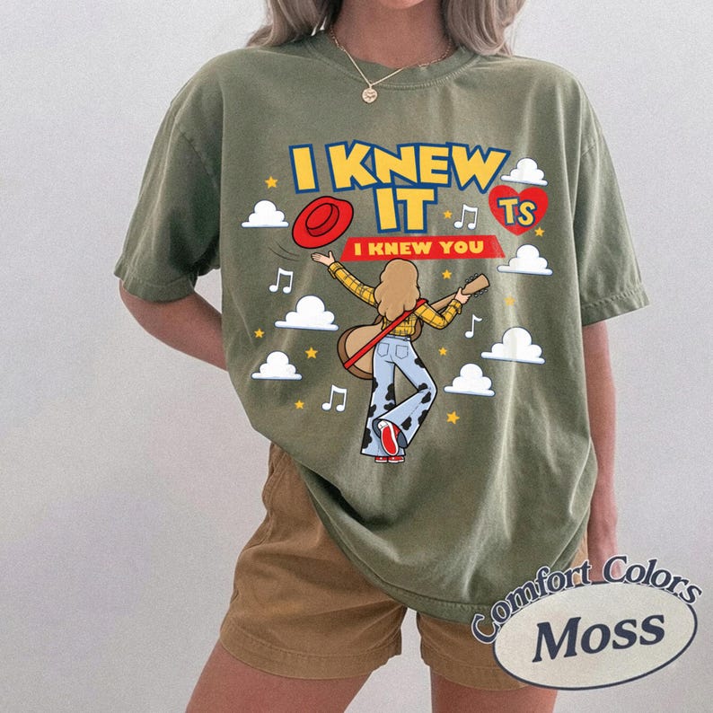 May include: An olive green t-shirt featuring a graphic print with the text I KNEW IT I KNEW YOU and TS inside a red heart. The design includes a cartoon figure with blonde hair wearing a yellow plaid shirt and cow print trousers, holding a guitar.