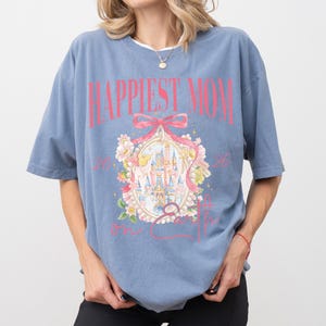 May include: A blue t-shirt with the text "Happiest Mom on Earth" in pink. The shirt features a castle graphic inside a decorative frame with floral accents. The shirt has short sleeves and a relaxed fit.