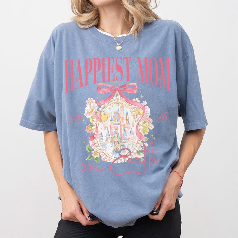 May include: A blue t-shirt with the text "Happiest Mom on Earth" in pink. The shirt features a castle graphic inside a decorative frame with floral accents. The shirt has short sleeves and a relaxed fit.