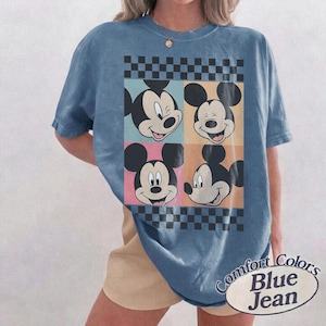 May include: Blue t-shirt featuring four panels with Mickey Mouse's face in different expressions. The panels are set against a checkered background. The shirt has the words "Comfort Colors Blue Jean" printed on it.
