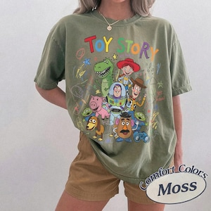 May include: Olive green t-shirt featuring a colorful graphic of Toy Story characters, including Buzz Lightyear, Woody, and Rex. The words "TOY STORY" are written in rainbow colors above the characters. The shirt is labeled "Comfort Colors Moss".