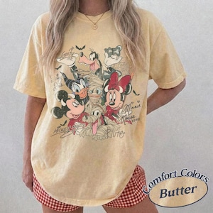 May include: Pale yellow Comfort Colors t-shirt with a Halloween-themed Disney character graphic. The design includes Mickey Mouse, Minnie Mouse, Donald Duck, Goofy, and Pluto. The tee has short sleeves and a loose fit.