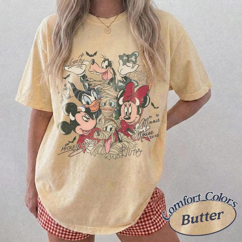 May include: Pale yellow Comfort Colors t-shirt with a Halloween-themed Disney character graphic. The design includes Mickey Mouse, Minnie Mouse, Donald Duck, Goofy, and Pluto. The tee has short sleeves and a loose fit.