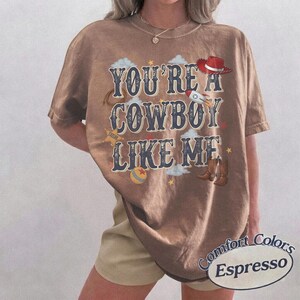 May include: A brown short-sleeve t-shirt featuring the text "YOU'RE A COWBOY LIKE ME" in dark blue lettering. The design includes illustrations of a red cowboy hat, a rocket, a ball, a lasso, and brown cowboy boots, accented with small stars and clouds.