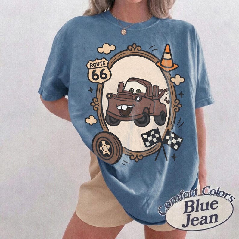 May include: Blue jean-colored t-shirt featuring a cartoon image of Mater from the movie Cars, framed by an ornate border. The design includes Route 66 signage, a traffic cone, and checkered flags. The shirt has the words "Comfort Colors Blue Jean" printed on it.