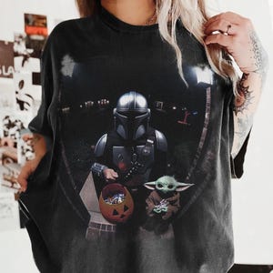 May include: Black t-shirt featuring a graphic of The Mandalorian and Grogu from the Star Wars series. The Mandalorian is holding a pumpkin-shaped candy bucket, and Grogu is holding a treat. The design is set against a nighttime backdrop.