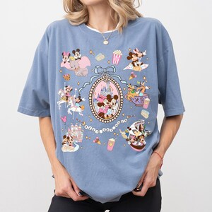 May include: Blue t-shirt featuring a colorful graphic of Disney characters, including Mickey and Minnie Mouse, in various scenes. The design includes a castle, ice cream, and popcorn. The shirt has short sleeves and a crew neck.