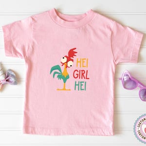 Peut inclure: T-shirt rose clair pour enfant avec un dessin de coq de dessin anim&eacute; et le texte "HEI GIRL HEI" en jaune, rouge et vert. Le coq a une cr&ecirc;te rouge, des plumes vertes et un bec et des pattes jaunes.