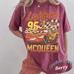 May include: Berry-colored t-shirt featuring a graphic of Lightning McQueen from the Cars movie. The design includes the number 95, a checkered flag, and the name "McQueen" in a vintage style. The shirt is labeled "Comfort Colors."