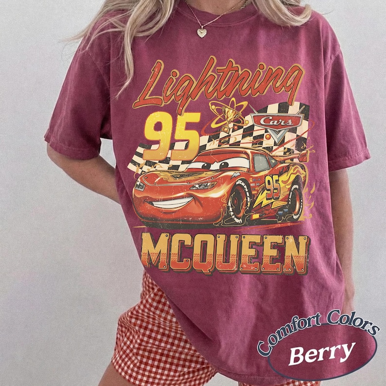 May include: Berry-colored t-shirt featuring a graphic of Lightning McQueen from the Cars movie. The design includes the number 95, a checkered flag, and the name "McQueen" in a vintage style. The shirt is labeled "Comfort Colors."