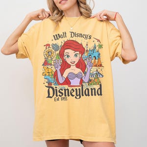 May include: Yellow t-shirt featuring a graphic of Ariel from The Little Mermaid, with the text "Walt Disney's Disneyland Est 1955". The design includes a Ferris wheel, castle, and other park elements.