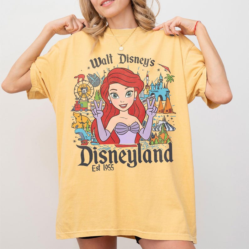 May include: Yellow t-shirt featuring a graphic of Ariel from The Little Mermaid, with the text "Walt Disney's Disneyland Est 1955". The design includes a Ferris wheel, castle, and other park elements.