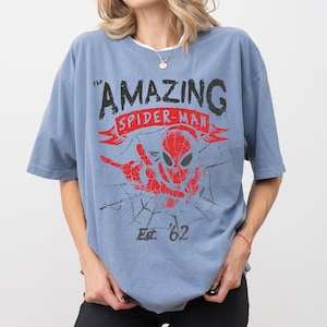 Pode incluir: Uma camiseta azul com as palavras "The Amazing Spider-Man" em preto e vermelho, com um gr&aacute;fico do rosto e da m&atilde;o do Homem-Aranha. A camisa tamb&eacute;m tem "Est. '62" impresso abaixo.
