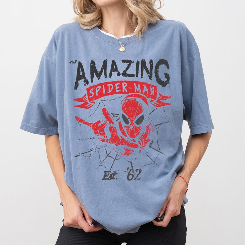 Pode incluir: Uma camiseta azul com as palavras "The Amazing Spider-Man" em preto e vermelho, com um gr&aacute;fico do rosto e da m&atilde;o do Homem-Aranha. A camisa tamb&eacute;m tem "Est. '62" impresso abaixo.