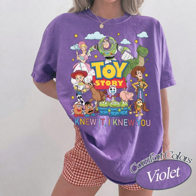 May include: A violet short-sleeved t-shirt featuring a colorful graphic print of Toy Story characters including Buzz Lightyear, Woody, Jessie, and others. The text "TOY STORY" is printed in large yellow letters, with "I KNEW IT, I KNEW YOU" below.
