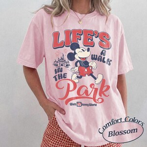 May include: A pale pink t-shirt featuring a Mickey Mouse graphic and the text "Life's a Walk in the Park" and "Walt Disney World". The shirt also has the words "Comfort Colors Blossom" printed on it.
