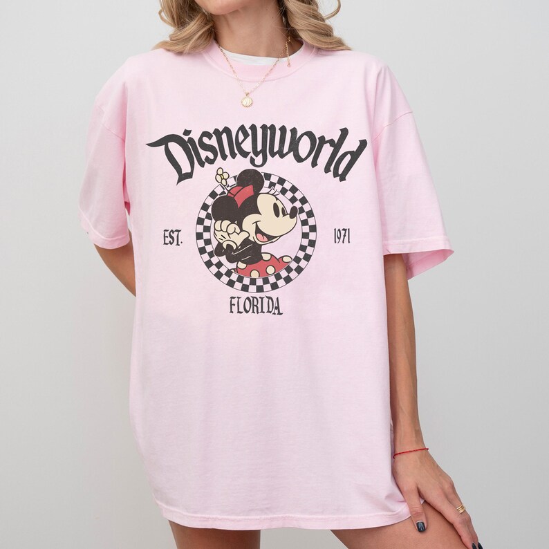 May include: A light pink t-shirt featuring the text "Disneyworld" in a script font. The shirt has a graphic of Minnie Mouse in a checkered circle, with "EST. 1971" and "FLORIDA" printed below.
