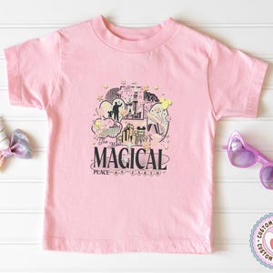 May include: A pale pink t-shirt with a graphic design that reads "MAGICAL PLACE ON EARTH". The design includes illustrations of a castle, Mickey Mouse, and other whimsical elements. The shirt is a short-sleeved crew neck.