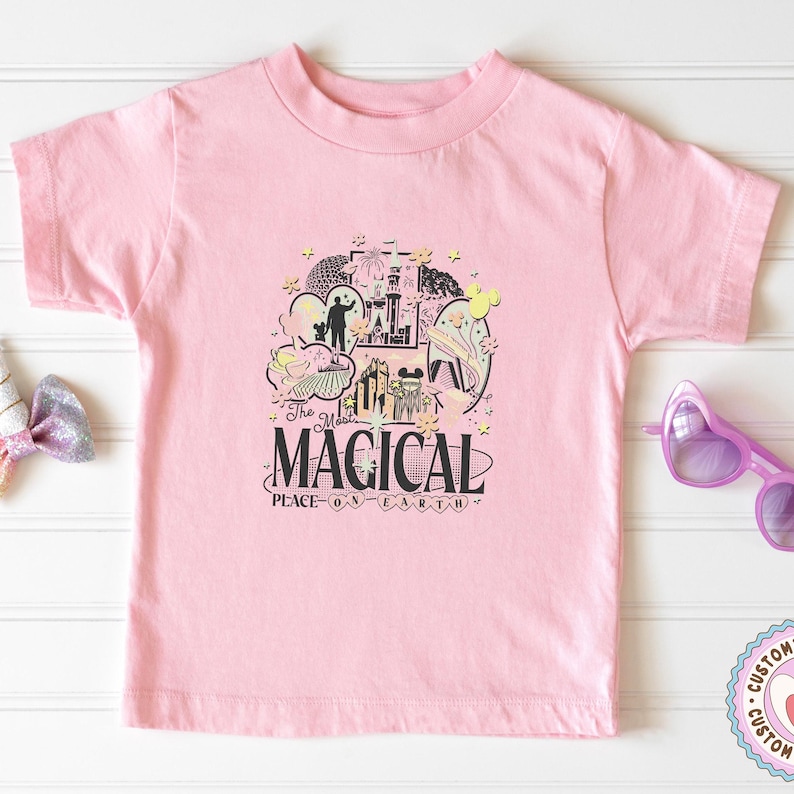 May include: A pale pink t-shirt with a graphic design that reads "MAGICAL PLACE ON EARTH". The design includes illustrations of a castle, Mickey Mouse, and other whimsical elements. The shirt is a short-sleeved crew neck.