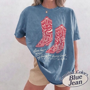 May include: A blue denim-coloured t-shirt featuring a graphic of two red cowboy boots decorated with white hearts, stars, and the number 13. Below the boots, the text reads, love has ways of bringing things back to life in white cursive script.