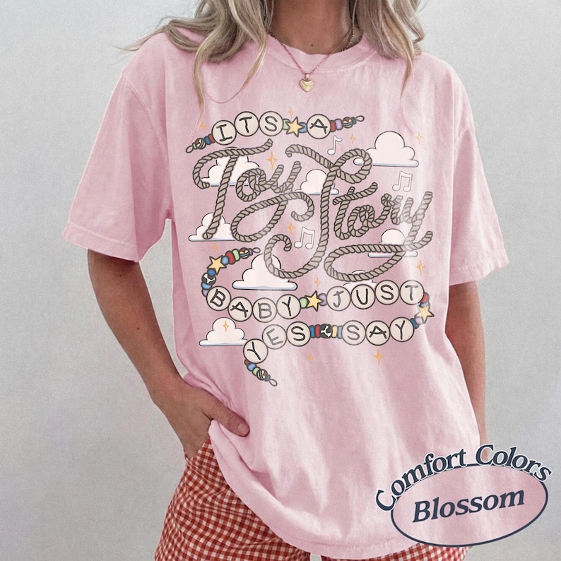 May include: A light pink short-sleeve t-shirt featuring a graphic print with the text "ITS A Toy Story BABY JUST YES SAY" written in rope-style lettering, surrounded by colorful beads, stars, musical notes, and white cloud illustrations.