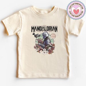 Może przedstawiać: Kremowa koszulka z grafiką The Mandalorian i Grogu otoczonych kwiatami. Tekst "THE MANDALORIAN" i "THIS IS THE WAY" jest wydrukowany nad i pod ilustracją. Okrągłe logo znajduje się w prawym g&oacute;rnym rogu.