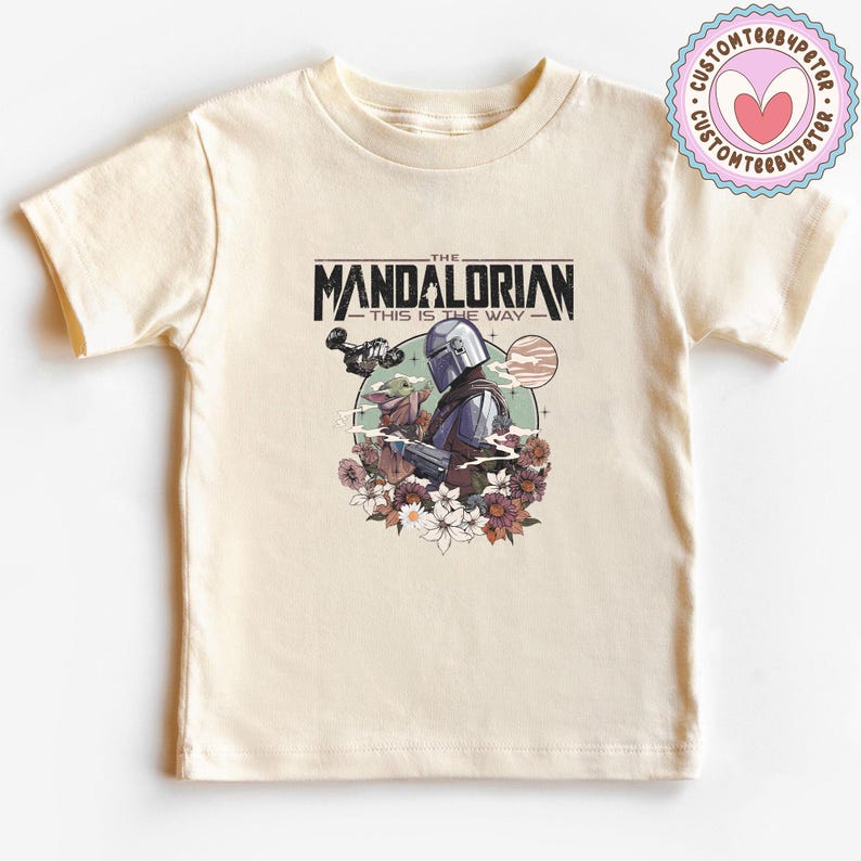 Może przedstawiać: Kremowa koszulka z grafiką The Mandalorian i Grogu otoczonych kwiatami. Tekst "THE MANDALORIAN" i "THIS IS THE WAY" jest wydrukowany nad i pod ilustracją. Okrągłe logo znajduje się w prawym g&oacute;rnym rogu.
