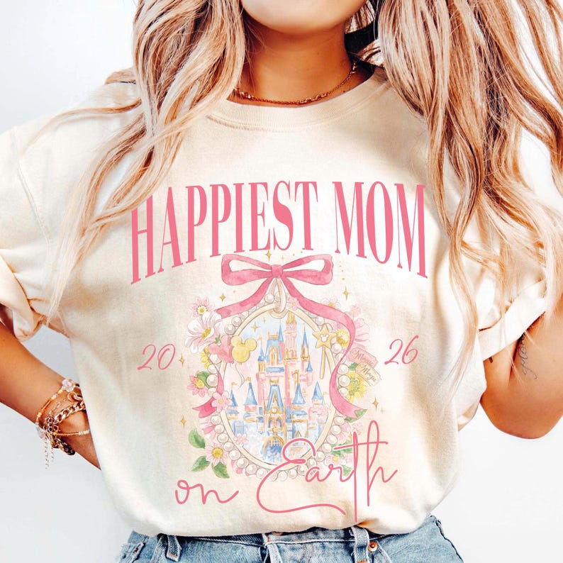 May include: Cream-colored sweatshirt with the text "Happiest Mom on Earth" in pink. The design includes a pink bow, a castle illustration, and the numbers "20" and "26". The sweatshirt has a crew neck.