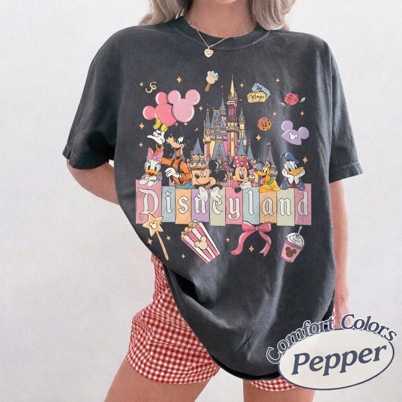 May include: Dark gray t-shirt featuring a colorful Disneyland graphic with Mickey Mouse, Minnie Mouse, Donald Duck, and other Disney characters. The design includes a castle, popcorn, and the word "Disneyland".