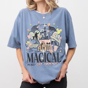 May include: A blue t-shirt with a graphic design featuring a castle, fireworks, and Mickey Mouse. The text on the shirt reads "The Most Magical Place on Earth" in a whimsical font, surrounded by hearts and stars.