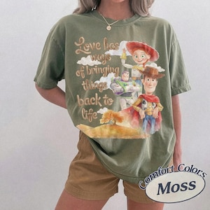 K&ouml;nnte beinhalten: Ein olivgr&uuml;nes Kurzarm-T-Shirt mit einem Grafikdruck von Woody, Buzz Lightyear und Jessie aus Toy Story. Der Text lautet, Love has ways of bringing things back to life, erg&auml;nzt durch Illustrationen von Wolken und einem kleinen Baum.