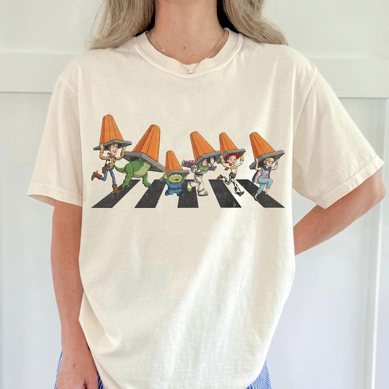 Puede incluir: Camiseta de color crema con un gr&aacute;fico l&uacute;dico de personajes animados cruzando una calle, cada uno con un cono de tr&aacute;fico naranja. El dise&ntilde;o es una parodia de una famosa portada de &aacute;lbum. Los personajes est&aacute;n en un paso de cebra en blanco y negro.