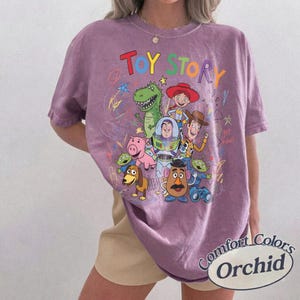 May include: A purple Comfort Colors t-shirt featuring a colorful graphic of Toy Story characters. The design includes the words "Toy Story" in rainbow colors, along with images of Woody, Buzz Lightyear, and other characters.