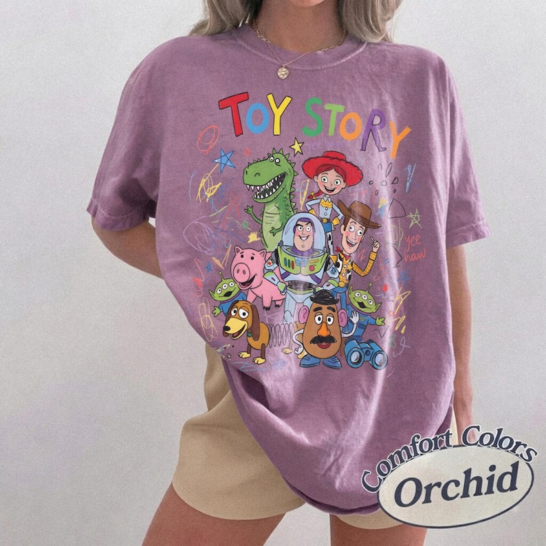 May include: A purple Comfort Colors t-shirt featuring a colorful graphic of Toy Story characters. The design includes the words "Toy Story" in rainbow colors, along with images of Woody, Buzz Lightyear, and other characters.