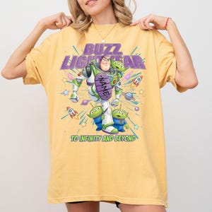 May include: A yellow t-shirt featuring a graphic of Buzz Lightyear and three green aliens. The text reads BUZZ LIGHTYEAR, NAME, and TO INFINITY AND BEYOND. The design includes colorful rockets, stars, and planets in a space-themed illustration.