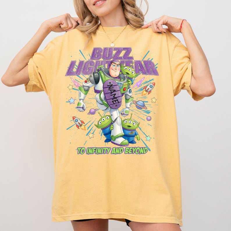 May include: A yellow t-shirt featuring a graphic of Buzz Lightyear and three green aliens. The text reads BUZZ LIGHTYEAR, NAME, and TO INFINITY AND BEYOND. The design includes colorful rockets, stars, and planets in a space-themed illustration.