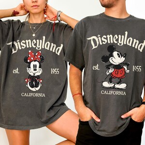 Może przedstawiać: Dwie szare koszulki z grafiką Disneyland. Jedna koszulka przedstawia Minnie Mouse z czerwoną kokardą, a druga Myszka Miki. Obie koszulki mają logo Disneyland, rok 1955 i napis "CALIFORNIA".