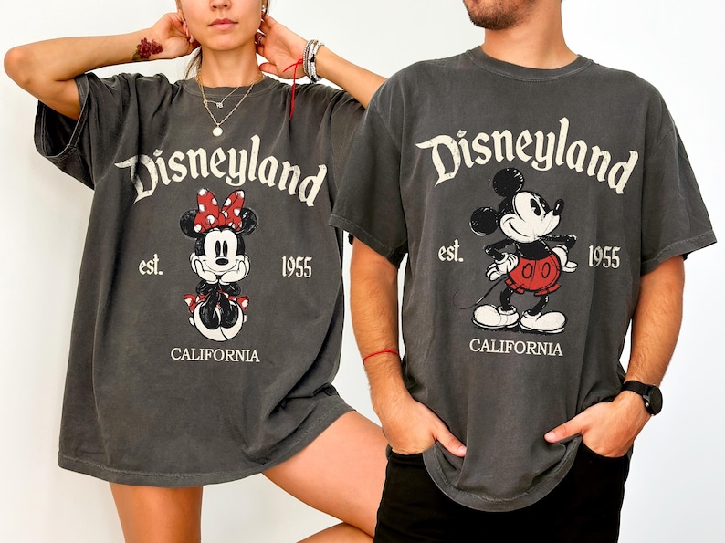Może przedstawiać: Dwie szare koszulki z grafiką Disneyland. Jedna koszulka przedstawia Minnie Mouse z czerwoną kokardą, a druga Myszka Miki. Obie koszulki mają logo Disneyland, rok 1955 i napis "CALIFORNIA".