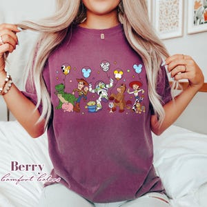 May include: Berry-colored t-shirt featuring a graphic of animated characters with balloons. The design includes a green dinosaur, a cowboy, a space ranger, a horse, and a cowgirl. The text "Berry Comfort Colors" is printed on the bottom left.