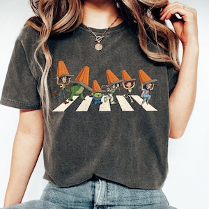 Puede incluir: Camiseta gris oscuro con una parodia de la portada del &aacute;lbum Abbey Road. El gr&aacute;fico muestra personajes de Toy Story cruzando un paso de cebra, cada uno con un cono de tr&aacute;fico naranja. Una camiseta divertida con tem&aacute;tica de cultura pop.