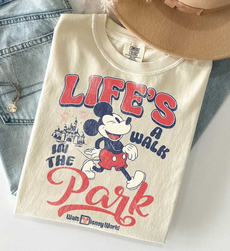 May include: Cream-coloured t-shirt featuring Mickey Mouse and the text "LIFE'S A WALK IN THE PARK" in red and blue. The design includes an illustration of Cinderella's castle and the words "Walt Disney World".