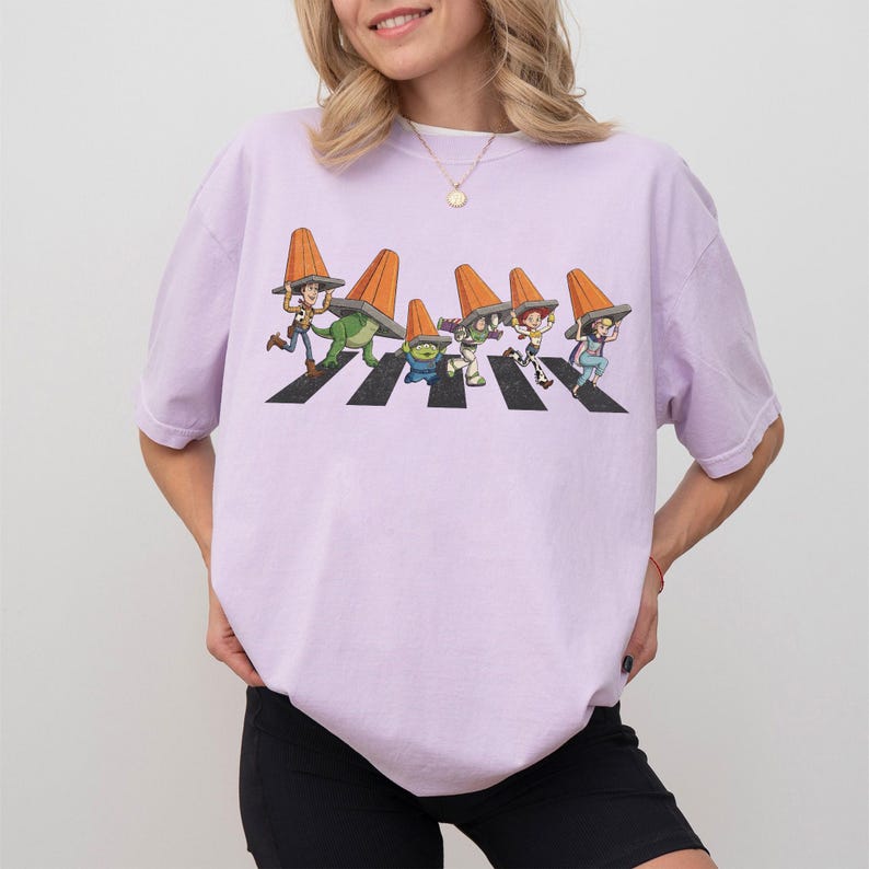 Puede incluir: Camiseta de color lila claro con un gr&aacute;fico de personajes de dibujos animados cruzando una calle, cada uno con un cono de tr&aacute;fico naranja. Los personajes est&aacute;n en un paso de cebra en blanco y negro. La camiseta es de corte holgado.