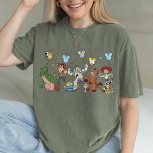 May include: Olive green t-shirt featuring a colorful graphic of cartoon characters holding balloons. The design includes a green dinosaur, a cowboy, a space ranger, a horse, and a cowgirl. The balloons have various patterns and shapes.
