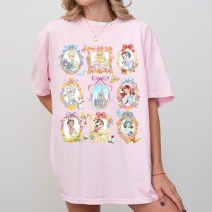 Pu&ograve; includere: T-shirt rosa chiaro con ritratti ad acquerello di principesse in cornici ornate. I ritratti sono circondati da motivi floreali e fiocchi. La maglietta ha maniche corte e scollo tondo.